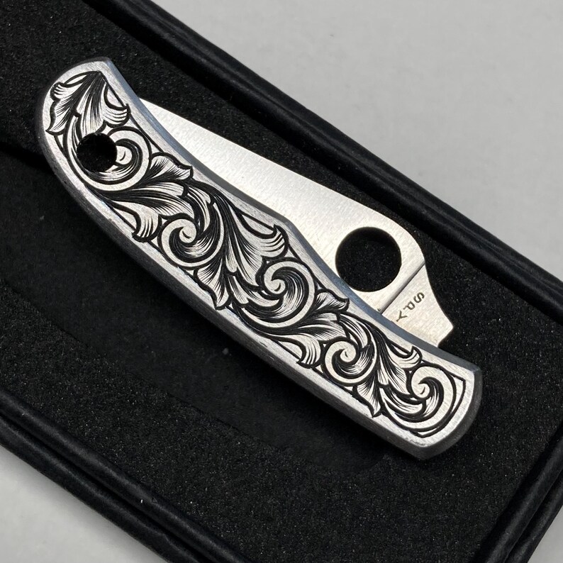Puede incluir: Una navaja plateada con un intrincado dise&ntilde;o floral negro grabado en el mango. La hoja est&aacute; parcialmente abierta. La navaja se presenta en una caja negra. El dise&ntilde;o es detallado.