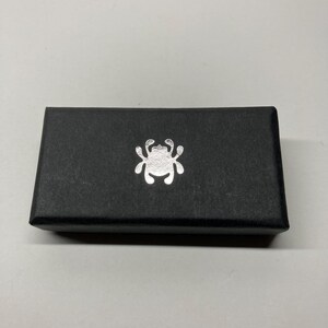 Puede incluir: Una caja rectangular negra con un emblema plateado en la parte delantera. El emblema es un dise&ntilde;o estilizado de insecto o ara&ntilde;a. La caja est&aacute; hecha de un material texturizado.