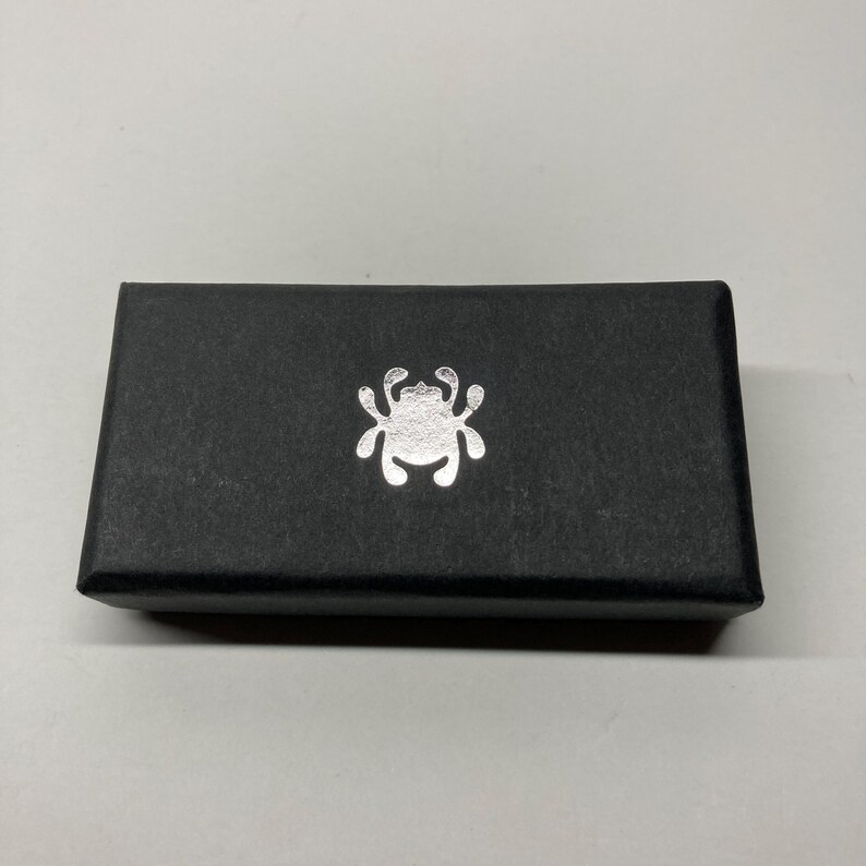 Puede incluir: Una caja rectangular negra con un emblema plateado en la parte delantera. El emblema es un dise&ntilde;o estilizado de insecto o ara&ntilde;a. La caja est&aacute; hecha de un material texturizado.