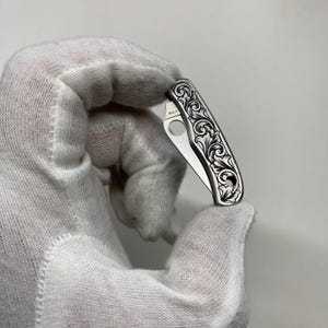 Puede incluir: Una peque&ntilde;a navaja plateada con un dise&ntilde;o floral ornamentado grabado en el mango. La hoja est&aacute; parcialmente abierta, revelando una peque&ntilde;a abertura circular. La navaja es sostenida por una mano enguantada sobre un fondo liso.