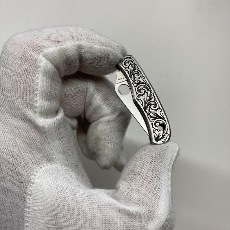 Puede incluir: Una peque&ntilde;a navaja plateada con un dise&ntilde;o floral ornamentado grabado en el mango. La hoja est&aacute; parcialmente abierta, revelando una peque&ntilde;a abertura circular. La navaja es sostenida por una mano enguantada sobre un fondo liso.