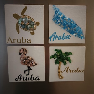 May include: Four square white tiles with Aruba-themed designs. One tile features a turtle, another a blue shape, a flamingo, and a palm tree. Each design is embellished with colorful, textured materials. The word "Aruba" is written on each tile.