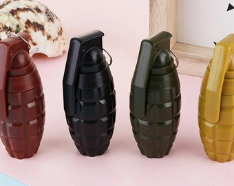 May include: Four toy grenade-shaped objects in a row, each in a different color: maroon, black, olive green, and mustard yellow. The grenades have a ring pull and a textured surface. A seashell is in the background.