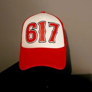 May include: A red and white trucker hat with the number "617" in bold, red lettering. The hat features a white front panel with the number and a red brim and mesh back. The hat is a classic baseball cap style.