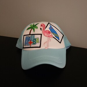 May include: A light blue trucker hat with a white front panel. The hat features a flamingo, palm tree, and the word "ARUBA" in a rectangular patch. A map of Aruba is also on the hat.