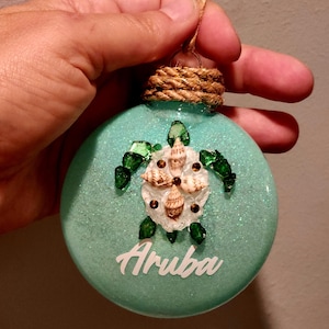 May include: A turquoise glittery Christmas ornament with a sea turtle design made of seashells and green glass pieces. The word "Aruba" is written in white script on the ornament. The ornament is suspended by a twine loop.