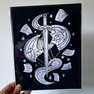 Puede incluir: Impresión artística en blanco y negro que representa una espada atravesando un libro, con líneas fluidas y páginas dispersas. El diseño incluye estrellas y elementos decorativos, creando un tema de fantasía. La impresión está en una funda de plástico transparente.
