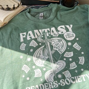 May include: A green sweatshirt with a white graphic design of a sword, book, and stars. The text "Fantasy Readers Society" and "Get Lost in a Book" are also included in the design.