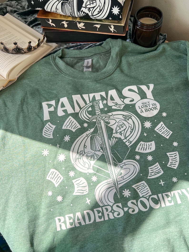 May include: A green sweatshirt with a white graphic design of a sword, book, and stars. The text "Fantasy Readers Society" and "Get Lost in a Book" are also included in the design.