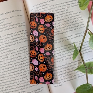 May include: A Halloween-themed bookmark featuring a black background with orange jack-o'-lanterns, pink leaves, and swirling vines. The bookmark is rectangular and is placed on top of an open book.