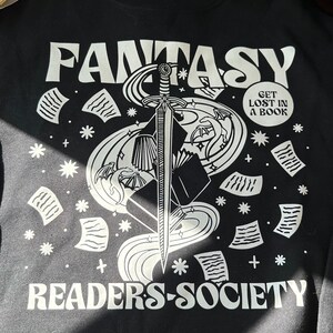 May include: Black long-sleeve shirt with a white graphic design of a sword, book, and dragon with the text "Fantasy Readers-Society" and a circle with the text "Get Lost In A Book".