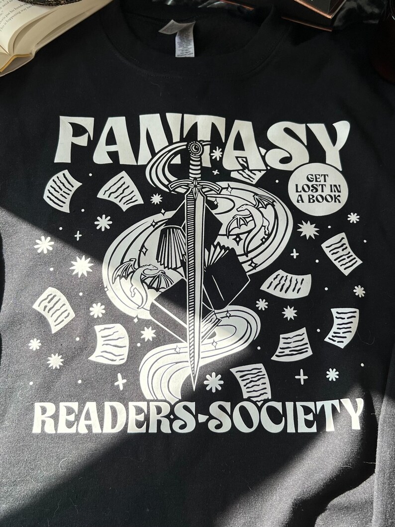 May include: Black long-sleeve shirt with a white graphic design of a sword, book, and dragon with the text "Fantasy Readers-Society" and a circle with the text "Get Lost In A Book".