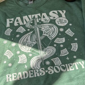 May include: A green t-shirt with a white graphic design of a sword, stars, and papers. The text "Fantasy Readers Society" and "Get Lost in a Book" are printed on the shirt.