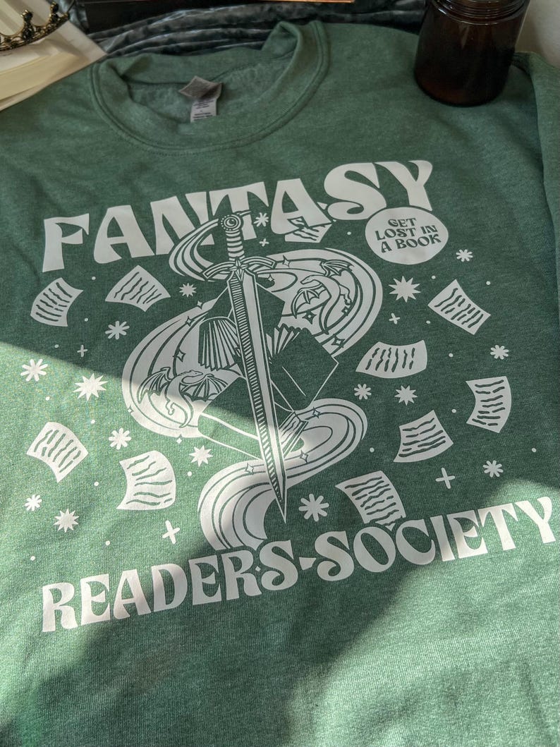 May include: A green t-shirt with a white graphic design of a sword, stars, and papers. The text "Fantasy Readers Society" and "Get Lost in a Book" are printed on the shirt.