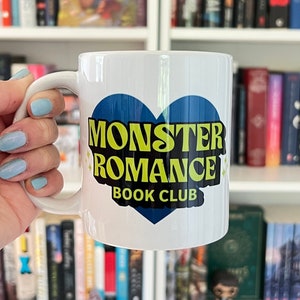 May include: White ceramic mug with a blue heart design. The heart has the text "Monster Romance Book Club" in yellow letters.