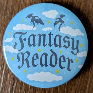 May include: A circular, light blue badge with the text "Fantasy Reader" in a decorative font. The badge has illustrations of dragons, clouds, and stars. The background is a light blue colour.