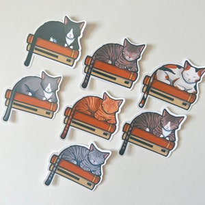 May include: Seven colorful cat stickers with different colored fur and patterns. Each cat is sleeping on top of a stack of books. The stickers are on a white background.