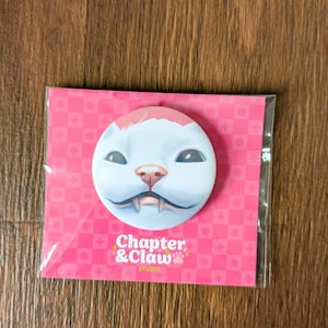 Puede incluir: Un pin redondo con la cara de un gato de dibujos animados en rosa y blanco, empaquetado en una tarjeta rosa. El texto "Chapter & Claw Studio" es visible. El fondo es una superficie de madera.