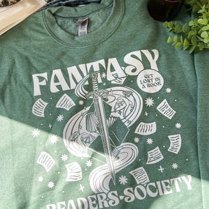 May include: A green sweatshirt with a white graphic design of a sword, book, and swirling smoke. The text "Fantasy Readers Society" and "Get Lost in a Book" are also included in the design.