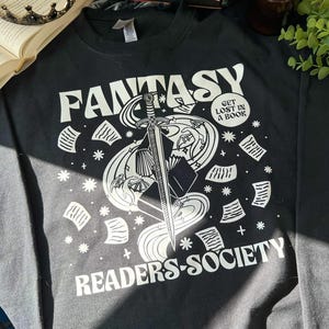 May include: Black sweatshirt with a white graphic design of a sword, book, and stars. The text "Fantasy Readers-Society" and "Get Lost In A Book" are also printed on the sweatshirt.