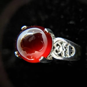 May include: A silver ring with a large, round, deep red gemstone. The ring's band features an openwork design with decorative cutouts. The gemstone is set in a four-prong setting, and the ring is displayed against a dark background.
