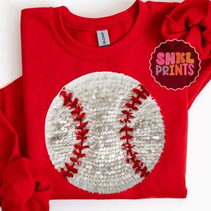 May include: A red sweatshirt featuring a large baseball design. The baseball is white with red stitching and is embellished with sequins. The sweatshirt has the brand name "Gildan" on the tag. The logo "SNKL PRINTS" is on the upper right.