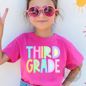 May include: A pink t-shirt with the words "THIRD GRADE" in colorful block letters. The person in the image is wearing pink sunglasses and making a peace sign with their fingers. The background has painted suns.
