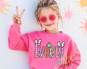May include: A pink sweatshirt with a colorful graphic of the name "Everly" with bunny ears and a green plant. The graphic is outlined in black and has a white polka dot pattern. The sweatshirt is made by Snkl Prints.