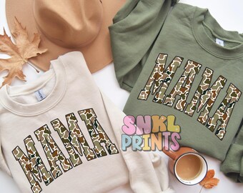 May include: Two crewneck sweatshirts, one beige and one olive green, each with the word "MAMA" in a camouflage pattern. A brown hat and a fall leaf are also visible. The sweatshirts are part of a clothing collection.