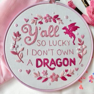 May include: A circular embroidery hoop containing white fabric with pink and magenta thread stitching. The text reads Y'all SO LUCKY I DON'T OWN A DRAGON, surrounded by embroidered pink flowers, leaves, small hearts, and a flying dragon.