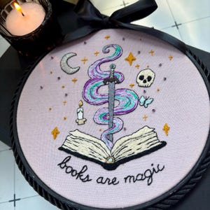 May include: A circular embroidery hoop featuring a light purple fabric background with a hand-stitched design of an open book, a sword, a crescent moon, a skull, and a candle. The text "books are magic" is stitched in black cursive below the book.