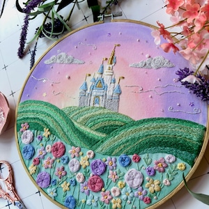 May include: A circular embroidery hoop featuring a hand-stitched scene of a castle with blue and gold turrets atop rolling green hills. The foreground is filled with colorful embroidered flowers in shades of pink, blue, and yellow against a sky.