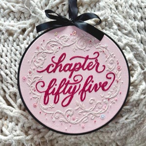 May include: A circular embroidery hoop featuring light pink fabric with the dark pink embroidered text "chapter fifty five" in a cursive script. The text is surrounded by a white floral vine pattern, small pink beads, and sequins, topped with a black ribbon.