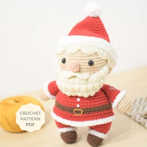 Pode incluir: Boneco de Papai Noel de crochê com chapéu vermelho, barba branca e botas marrons. O boneco está vestindo um terno vermelho e branco com um cinto marrom. O boneco está em pé sobre uma superfície de madeira. O texto "CROCHET PATTERN PDF" é visível na imagem.