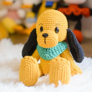 Pode incluir: Um brinquedo de cachorro de crochê amarelo com orelhas pretas e uma echarpe verde. O cachorro está sentado em uma superfície branca e fofa.