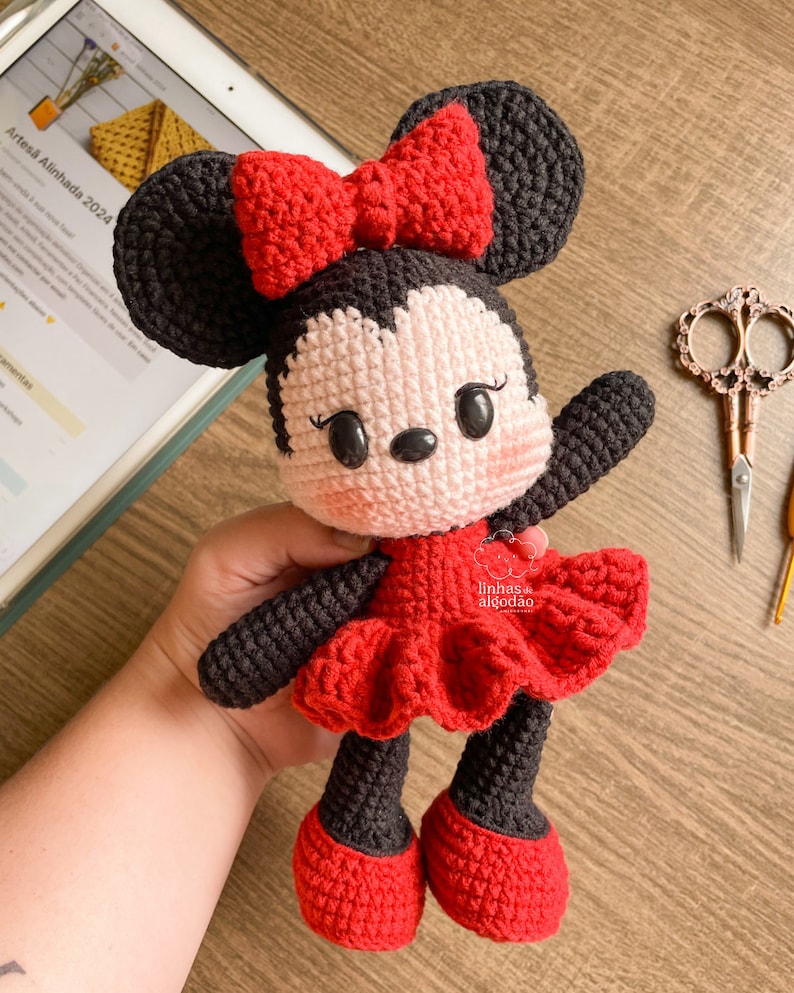 May include: A handmade crochet doll resembling a cartoon character. The doll has a black head with large ears, a red bow, and a red dress. The doll's face is light pink with black eyes and a small nose. The doll is holding its arm up.