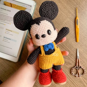 Puede incluir: Un muñeco de Mickey Mouse de crochet que lleva un mono amarillo con tirantes azules y una pajarita azul. El muñeco tiene orejas negras, una cara rosa y zapatos rojos. El muñeco está siendo sostenido por la mano de una persona.