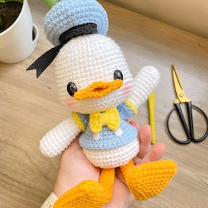 Pode incluir: Um pato de pelúcia de crochê vestindo um chapéu de marinheiro azul e preto, uma gravata borboleta amarela e uma camisa azul e branca. O pato tem um corpo branco, um bico laranja e pés amarelos.