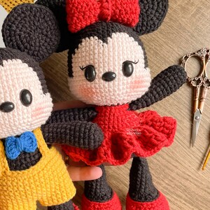 May include: Crocheted Minnie Mouse doll with a red dress and bow. The doll has black ears, arms, and legs, and a white face with rosy cheeks. A second doll, Mickey Mouse, is partially visible.