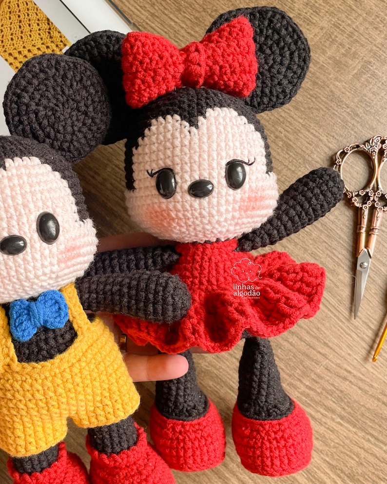 May include: Crocheted Minnie Mouse doll with a red dress and bow. The doll has black ears, arms, and legs, and a white face with rosy cheeks. A second doll, Mickey Mouse, is partially visible.