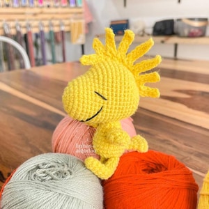 May include: A yellow crocheted cartoon character with a spiky yellow hairdo sitting on top of three balls of yarn. The yarn is in shades of grey, pink, and orange.