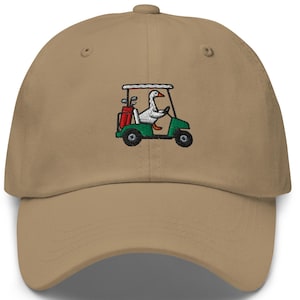 May include: A tan baseball cap with an embroidered design of a goose driving a green golf cart. The golf cart has a red golf bag in the back. The goose is wearing a white hat and is holding the steering wheel.