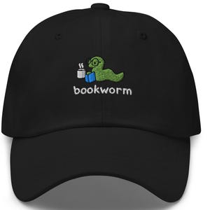 Puede incluir: Gorra de béisbol negra con un diseño bordado de un gusano de biblioteca verde. El gusano lleva gafas, un libro y una taza de café. La palabra "bookworm" está bordada debajo.