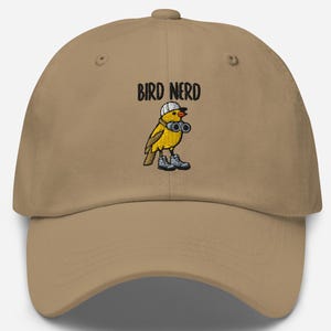 May include: A tan baseball cap with the embroidered text "BIRD NERD" above a cartoon bird wearing a hat, binoculars, and boots. The bird is yellow with black and white details.