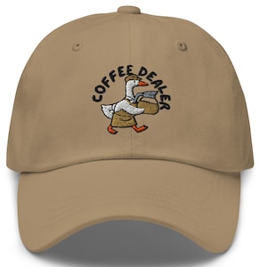 以下が含まれることがあります： ベージュ色の野球帽。つばがカーブしています。帽子には、コーヒーバッグを運ぶガチョウの刺繍デザインがあり、ガチョウの上に「COFFEE DEALER」という言葉が円形パターンで配置されています。