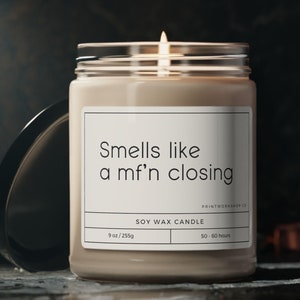 May include: A soy wax candle in a clear glass jar with a beige label that reads "Smells like a mf'n closing" and "SOY WAX CANDLE". The candle is 9 oz / 255g and has a burn time of 50-60 hours.