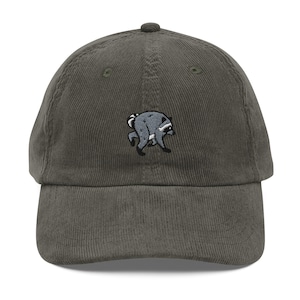 Op de afbeelding: Een donkergrijze corduroy baseballpet met een geborduurde afbeelding van een kleine wasbeer in een gebogen loophouding. De pet heeft een gestructureerde kroon, een gebogen klep en een knoop aan de bovenzijde van de pet.