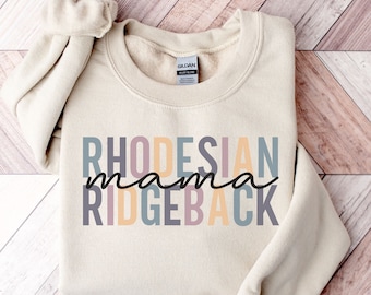 Puede incluir: Una sudadera beige con el texto "Rhodesian mama Ridgeback" en letras de colores. El texto está apilado verticalmente con "mama" en una fuente más pequeña y en negro.