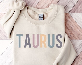Puede incluir: Una sudadera beige con la palabra "TAURUS" impresa en letras de colores. Las letras son en tonos de azul, rosa, amarillo y gris.