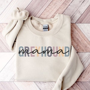 May include: A beige sweatshirt with the text "Greyhound mama" in colorful letters. The letters are in shades of pink, yellow, blue, and gray.